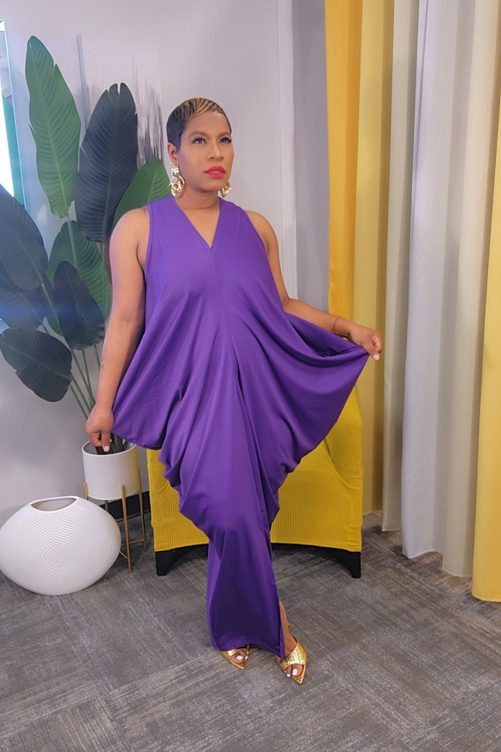 Woman in a purple dress posing in an indoor setting with yellow curtains and a plant.